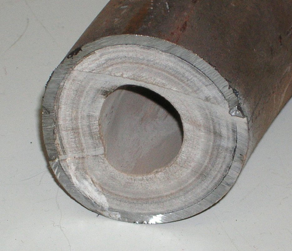 Cross-section of a cut metal pipe whose inside wall is lined with a thick, layered grey-white mineral crust.