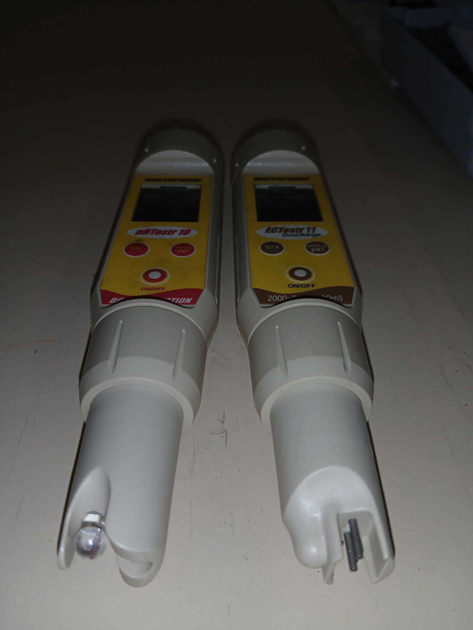 Two yellow and white pen-style testers side by side, caps off; one has a glass bulb electrode, the other two metal pins.