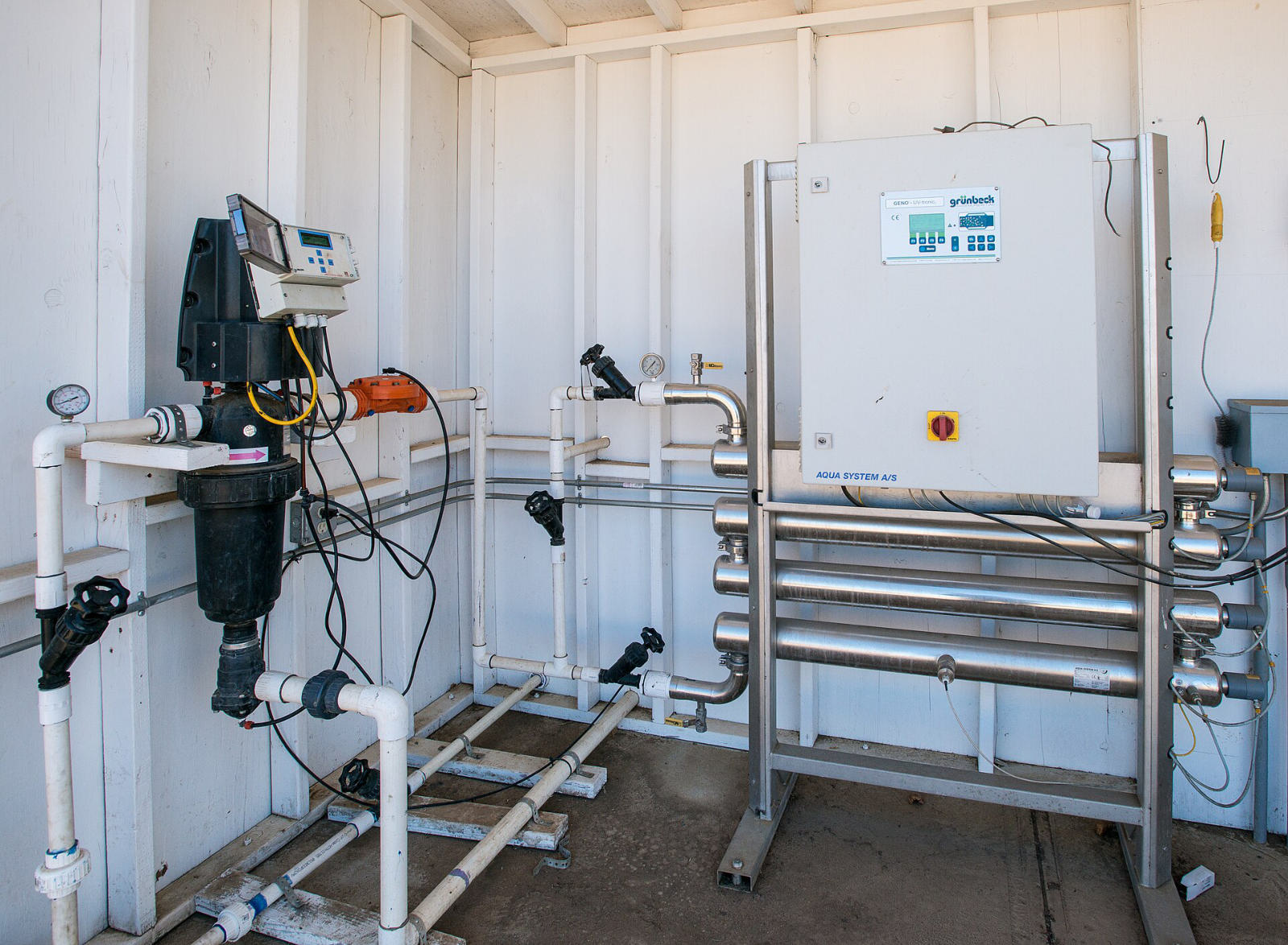 Inside a shed: a black filter with a controller on white plastic pipework, and a stainless steel ultraviolet steriliser with a control cabinet.