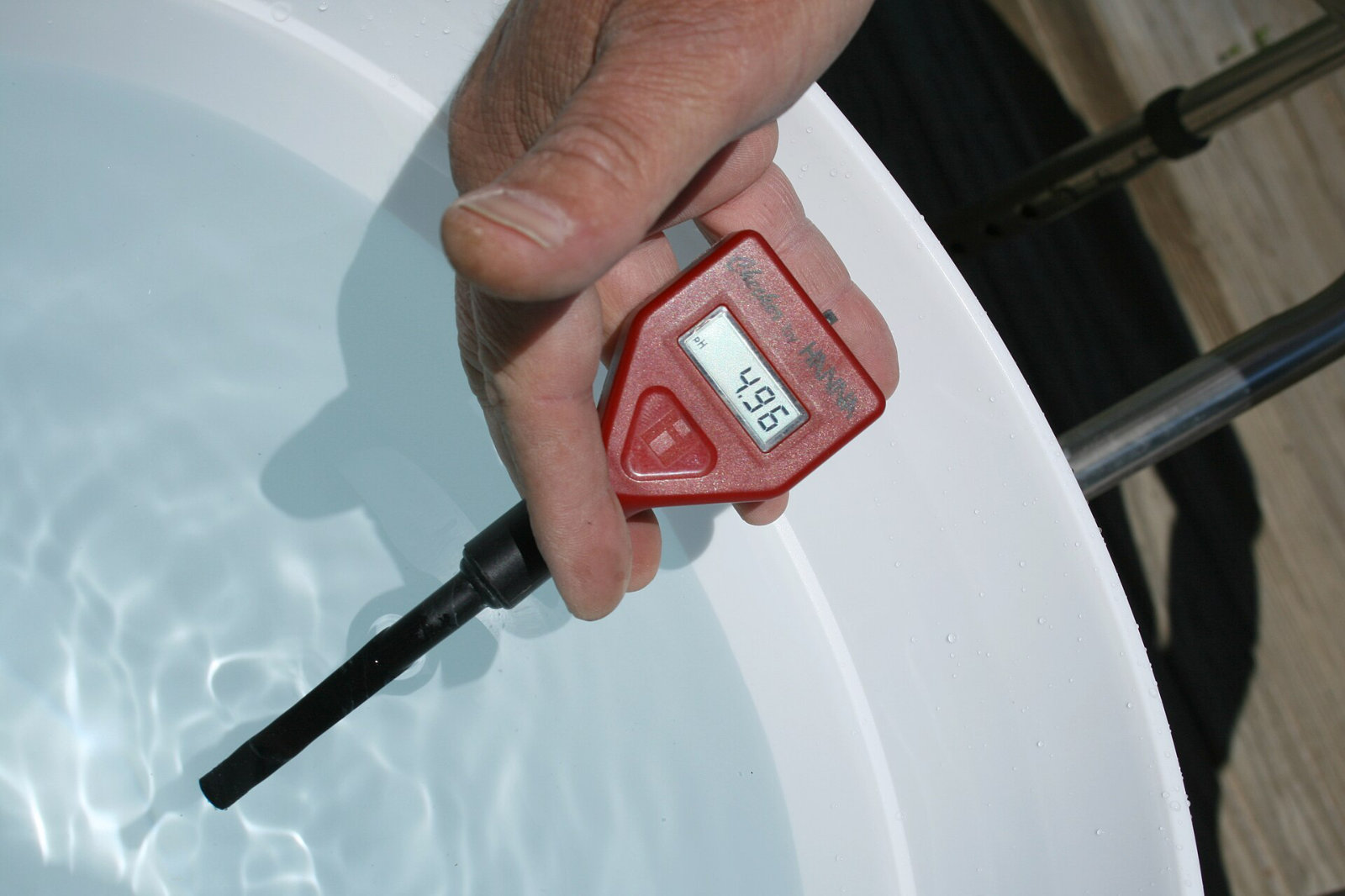 A hand holds a small red handheld pH meter with its probe in water in a white tub; the display reads 4.96.